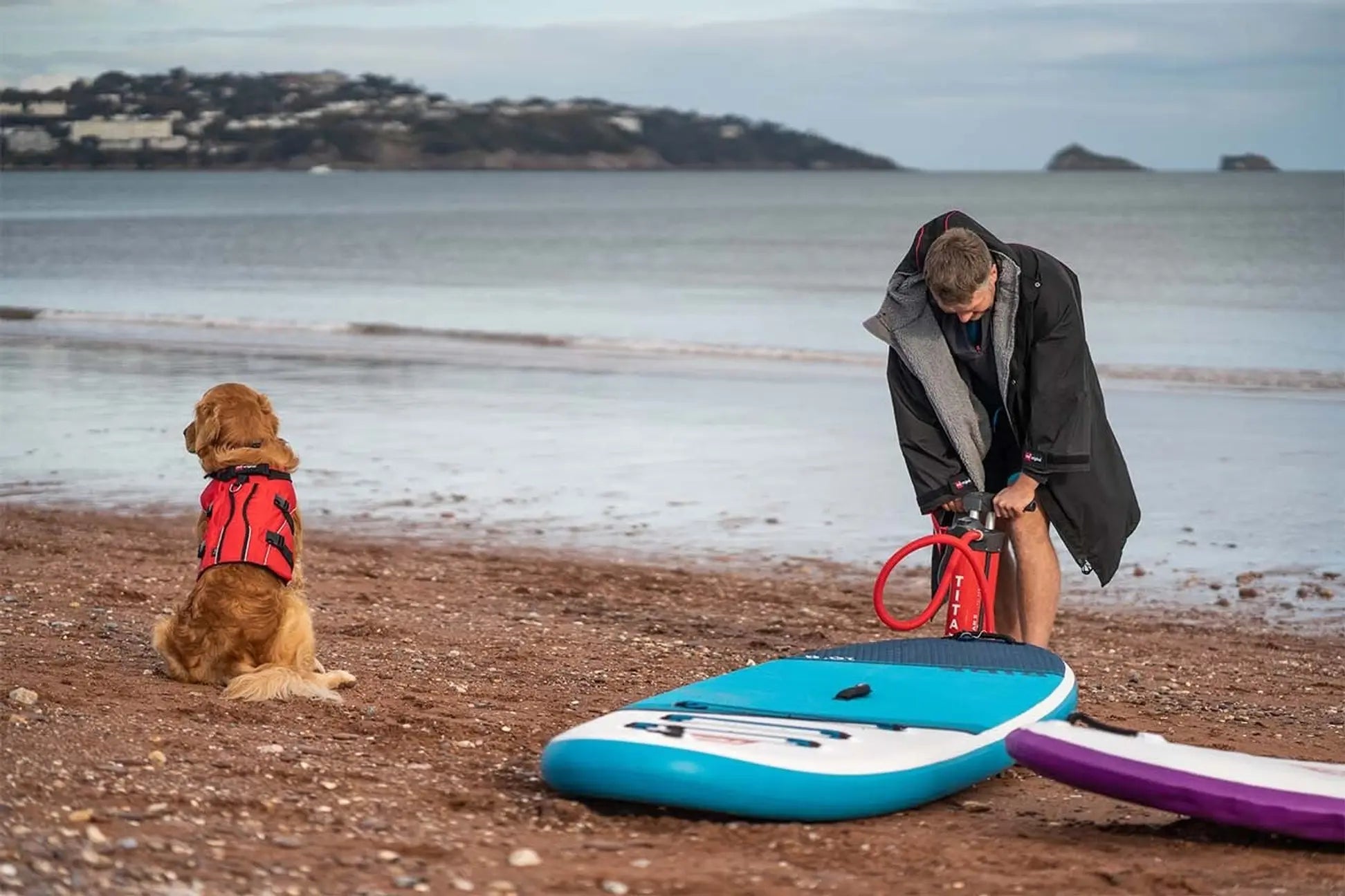 A man and his dog on the beach while he pumps up his paddleboard