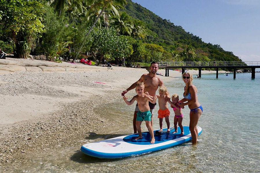 Family of five stood on a paddleboard in the shallows of the sea, camper van next to river with paddle boarder