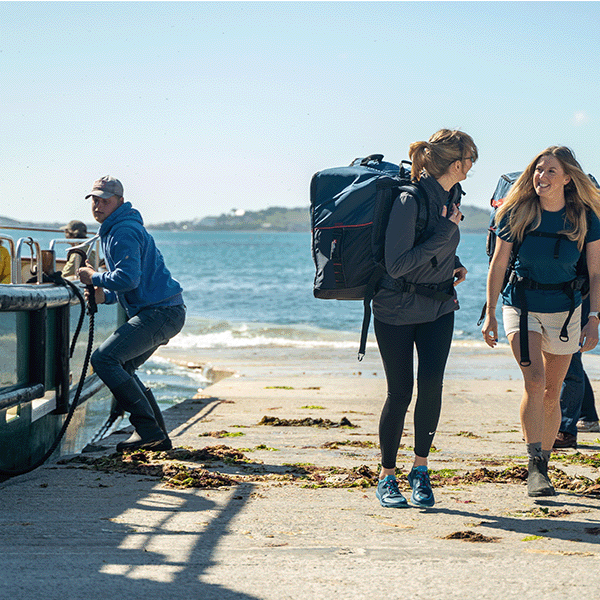 Two women walking up a slipway from a passenger ferry carrying Red Paddle Co Compact Inflatable Paddle Boards 