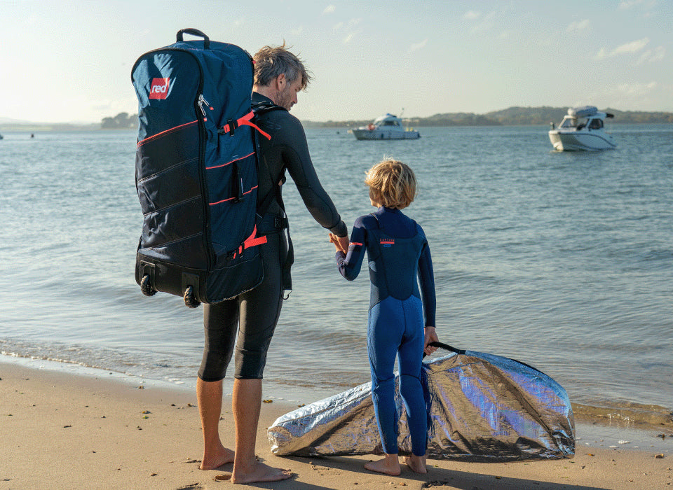 A man holding his sons hands walking along the beach wearing a backpack