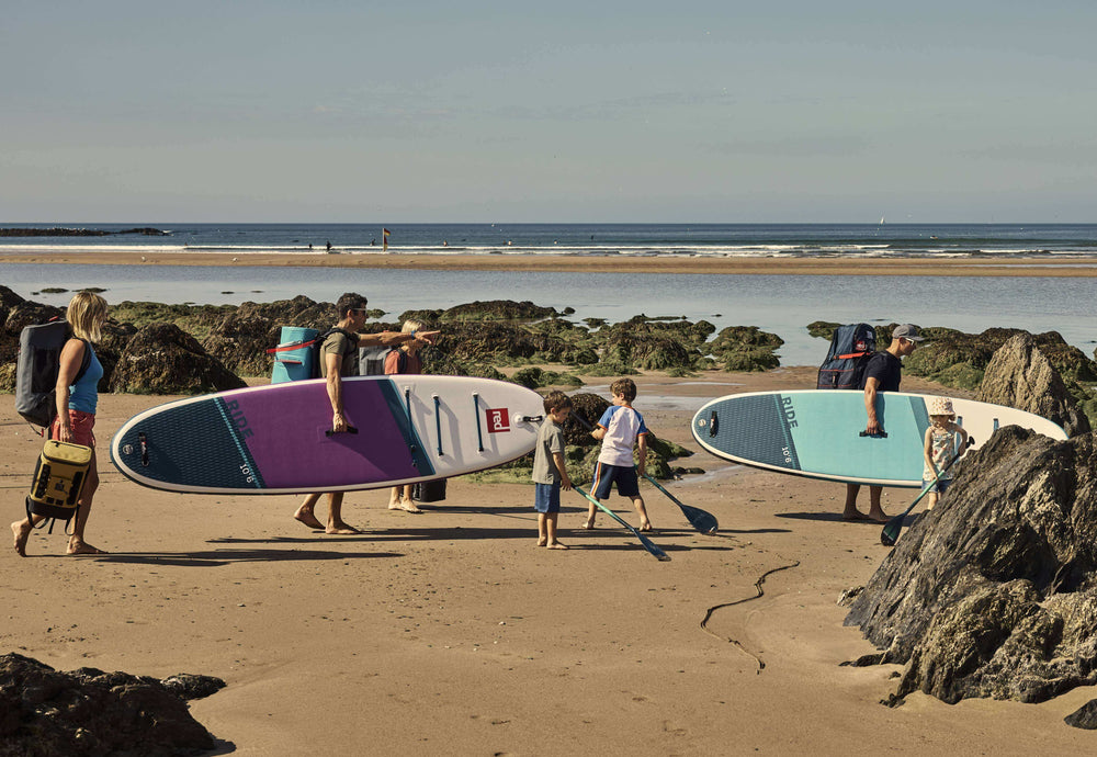 a family walking across the beach with red paddle boards, cooler bags