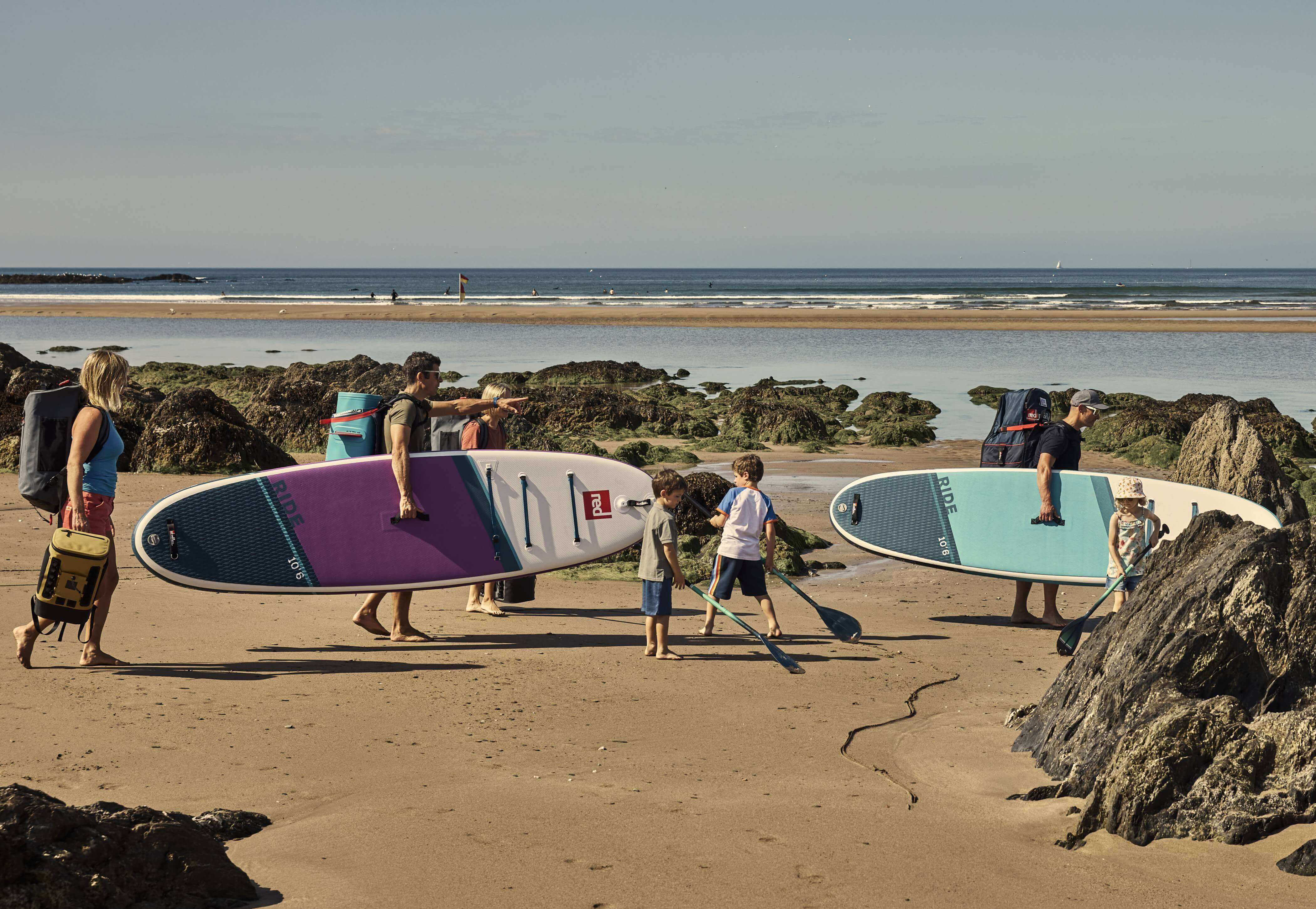 a family walking across the beach with red paddle boards, cooler bags