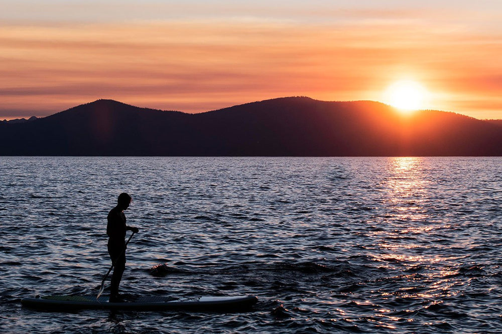 paddle boarder at sunset, lake surrounded by trees, silhouette on girl on SUP next to canyon, man jumps off rock with SUP on water below, Girl stands on paddle board on glassy still lake, girl paddles with mountains on the horizon