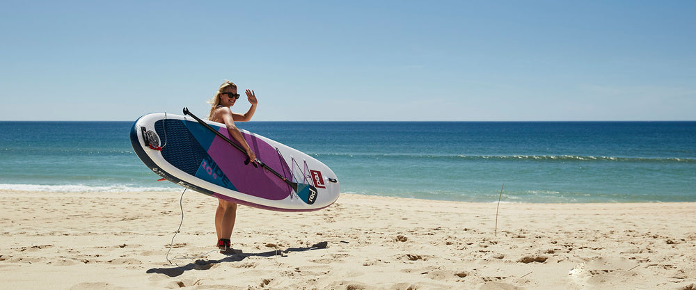 woman holding red paddleboard on the beach and waving to the camera