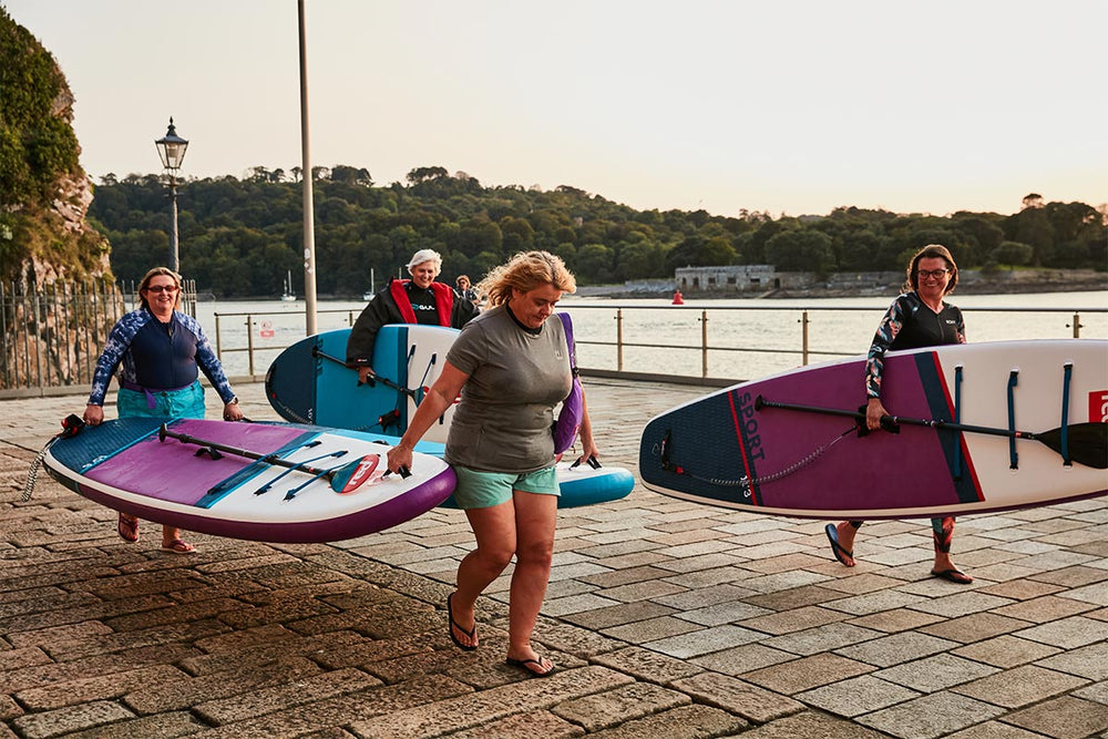 4 people carrying 4 paddle boards along a path by the sea