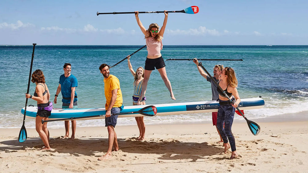 A group of people carrying a multiple person sized paddleboard along the beach