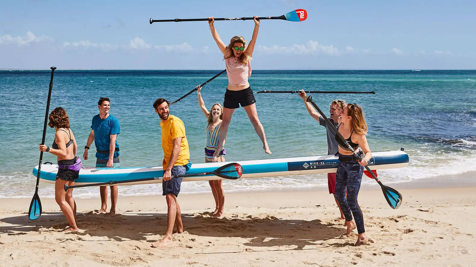 A group of people carrying a multiple person sized paddleboard along the beach