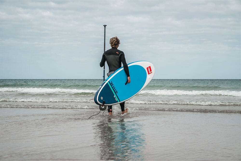 man wearing a wetsuit walking into the sea carrying the Whip MSL inflatable paddle board