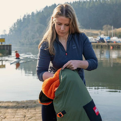 Woman stood next to river with a rower on a rowing boat in the background, wearing Red Original Performance Top Layer and putting a Pro Change Robe EVO in Parker Green into a Pro Change Stash Bag