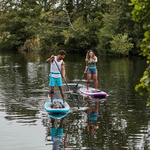A man and a woman stand up paddleboarding on a river amongst the trees featuring the Red Paddle Co 10'8 Ride Blue and 10'6 Ride Purple