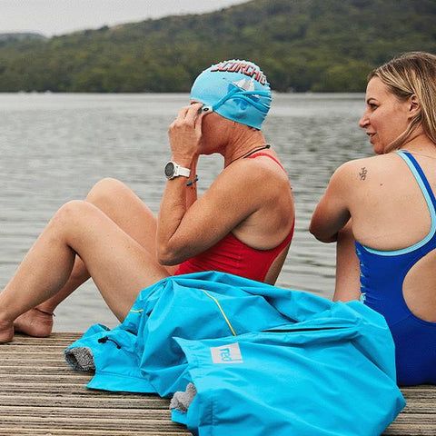Two women wearing swimsuits sat on a deck next to a lake before they swim - Red Original Pro Change Robe EVO in the foreground