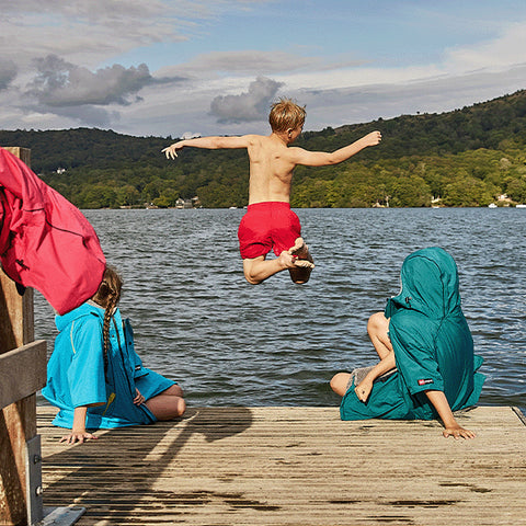 2 Children on a deck next to Lake Windemere wearing Red Original Pro Change Robe EVO, 1 child jumping into the water