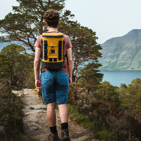 Man hiking on a footpath with a lake and mountains ahead of him, carrying a Red Original Waterproof Cool Bag Backpack in Mustard