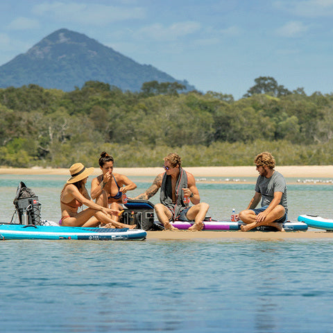 A group of friends sat on the beach with a Red Original Deck Bag, a Cool Bag, a Microfibre Towel and Ride Inflatable Paddle Boards
