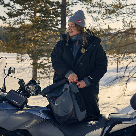Woman in the snow and the trees wearing Red Original Pro Change Robe EVO Swim Parka and Voyager Beanie in Graphite, holding the Waterproof Backpack and Waterproof Dry Pouch on the back of a snowmobile