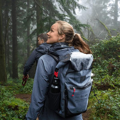 Man and woman walking through the mist and trees in the green woods wearing Red Original Performance Clothing Active Jacket, Waterproof Backpack and Stainless Steel Water Bottle in Black