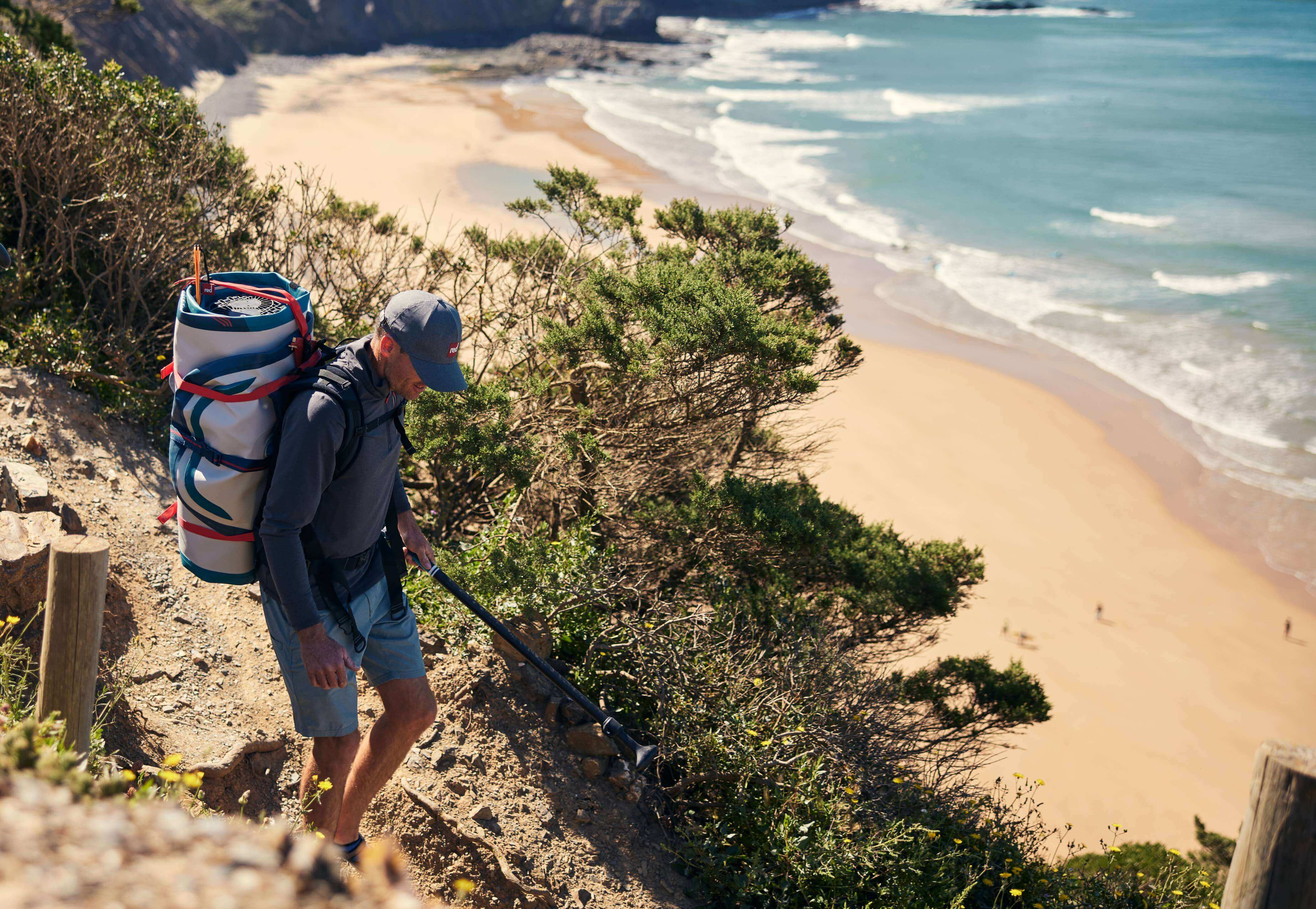 Man walking down a beach path, carrying a paddle board using Red's ATB transformer bag.