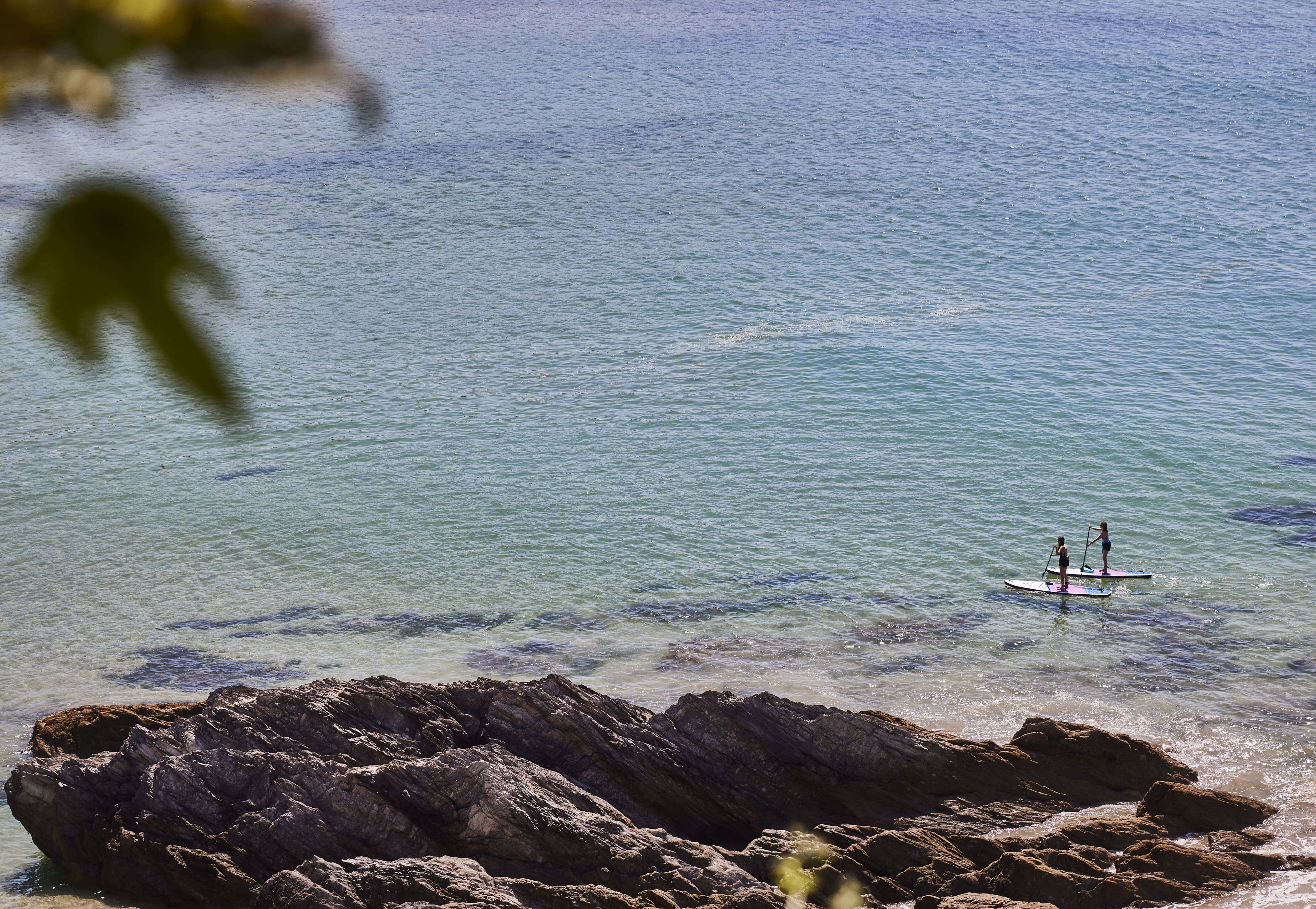 Two people on Red paddle boards, paddling around rocks on the beach.