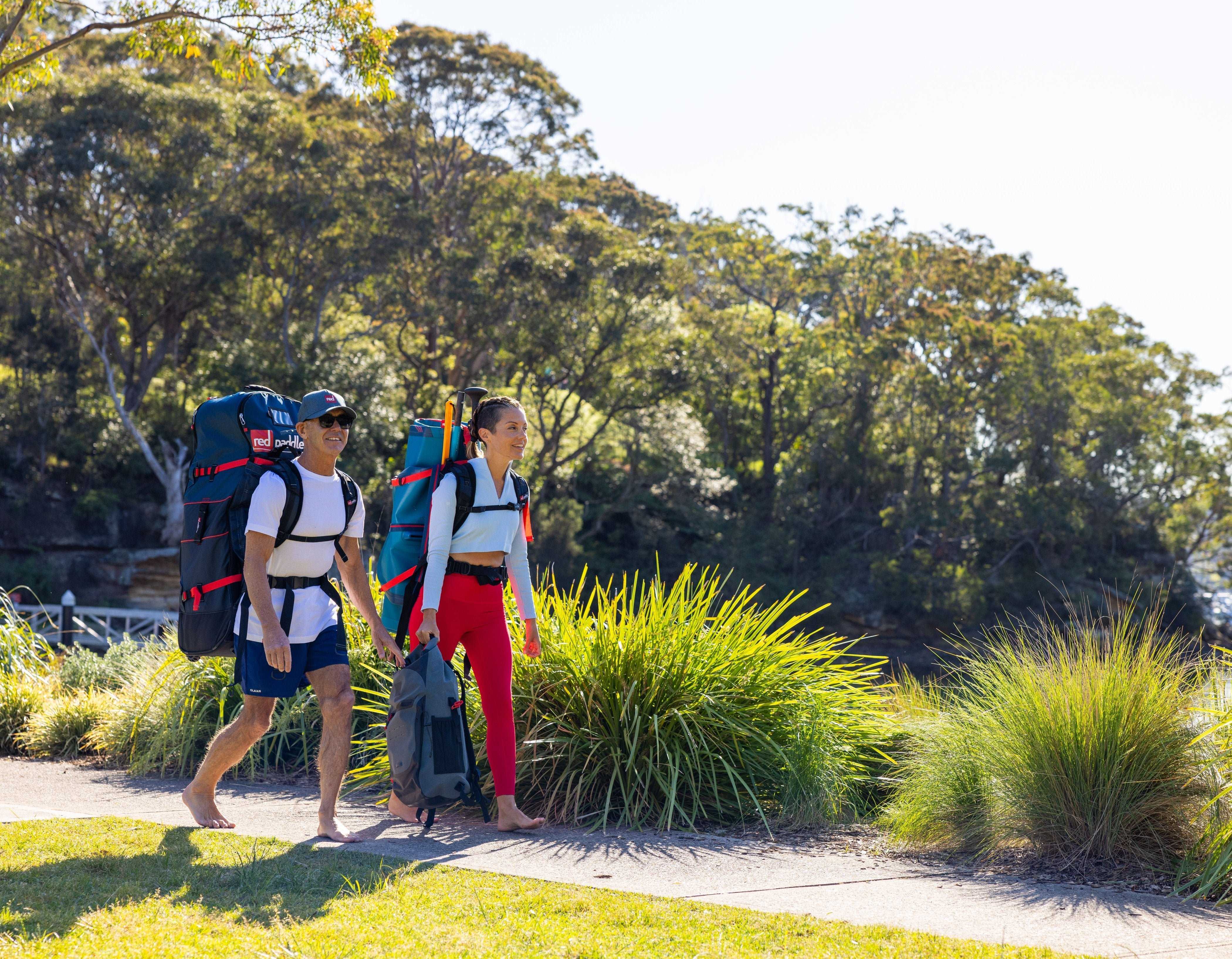 Two people walking beside a lake, carrying Red paddle boards using the ATB Transformer Board bag as well as carrying a kit bag.