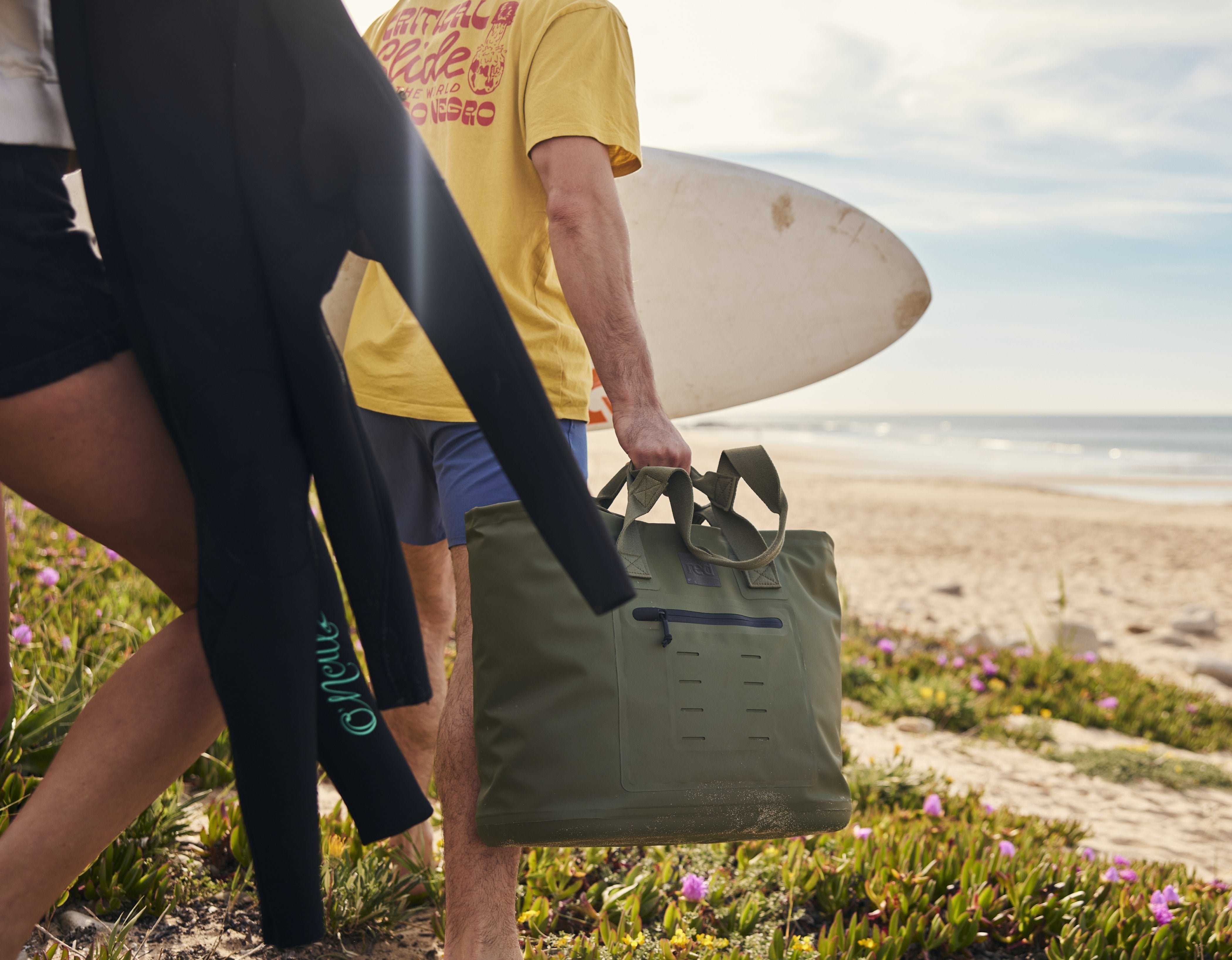 Two people walking onto the beach carrying a wetsuit and a Red Olive Green Waterproof Tote Bag. 